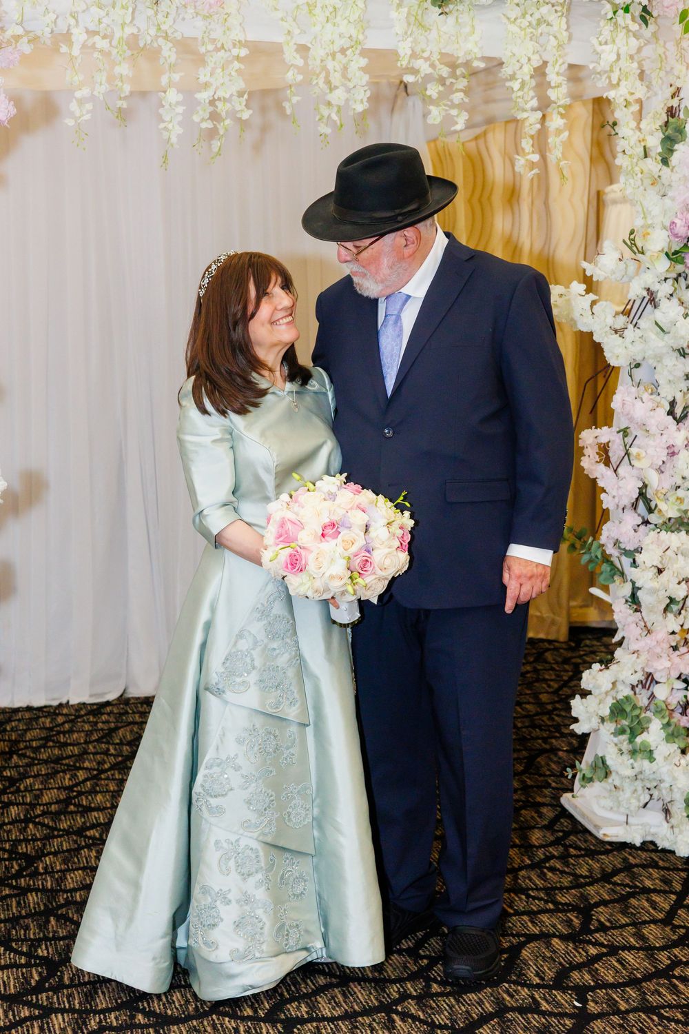 Elegant couple in wedding attire, woman in mint gown with bouquet, man in navy suit and black hat.