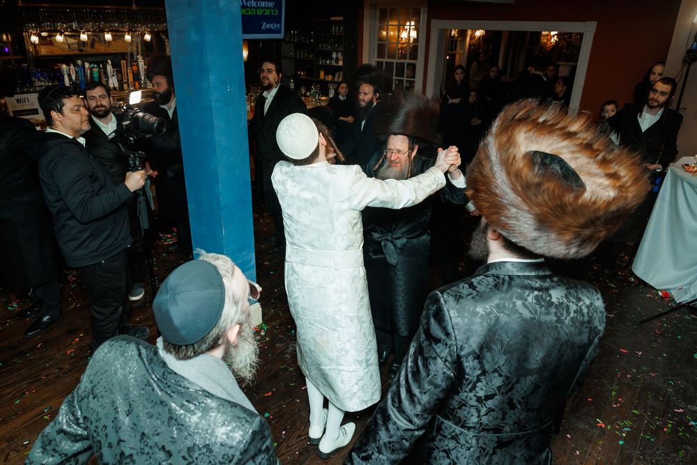 Jewish men dancing joyfully at a traditional celebration in a crowded indoor venue.