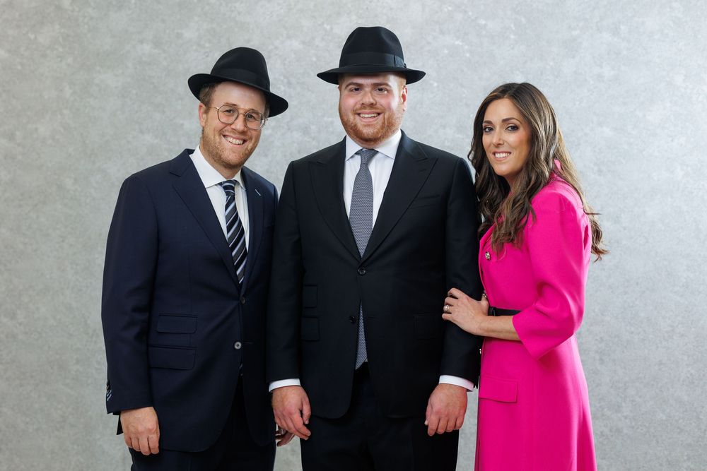 Two men in black suits and hats pose with a woman in a pink dress for a formal portrait photo.