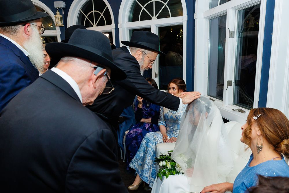 Rabbi blesses bride in white veil at traditional Jewish wedding ceremony with guests in black hats.
