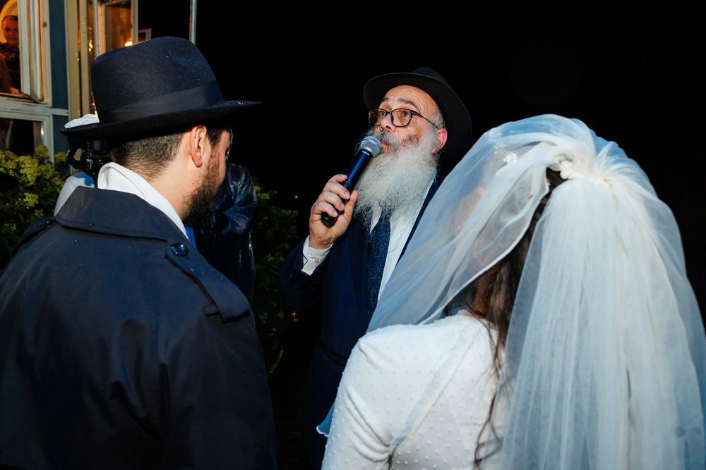 Rabbi officiating a Jewish wedding ceremony, speaking into microphone with bride in veil and groom in traditional hat.