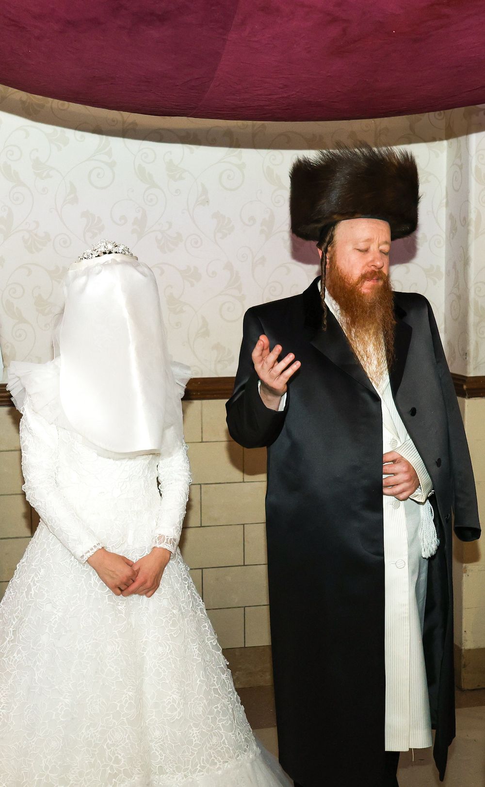 Orthodox Jewish groom in traditional black coat and fur shtreimel hat standing beside veiled bride in white wedding dress.