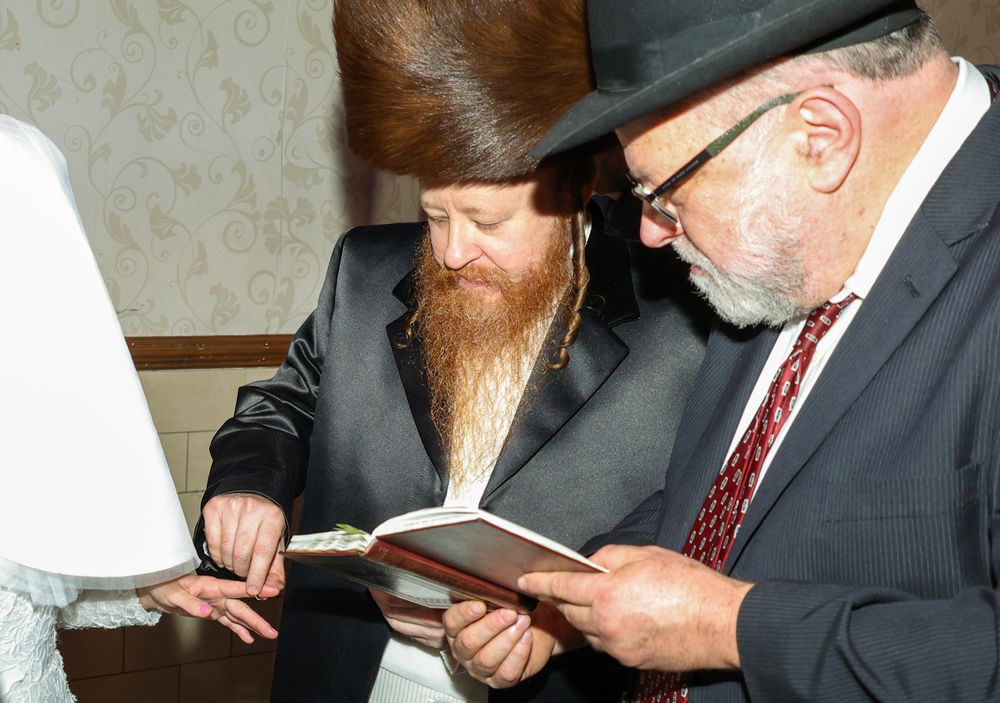 Two men in traditional Jewish attire reading from a book together at a formal indoor gathering.