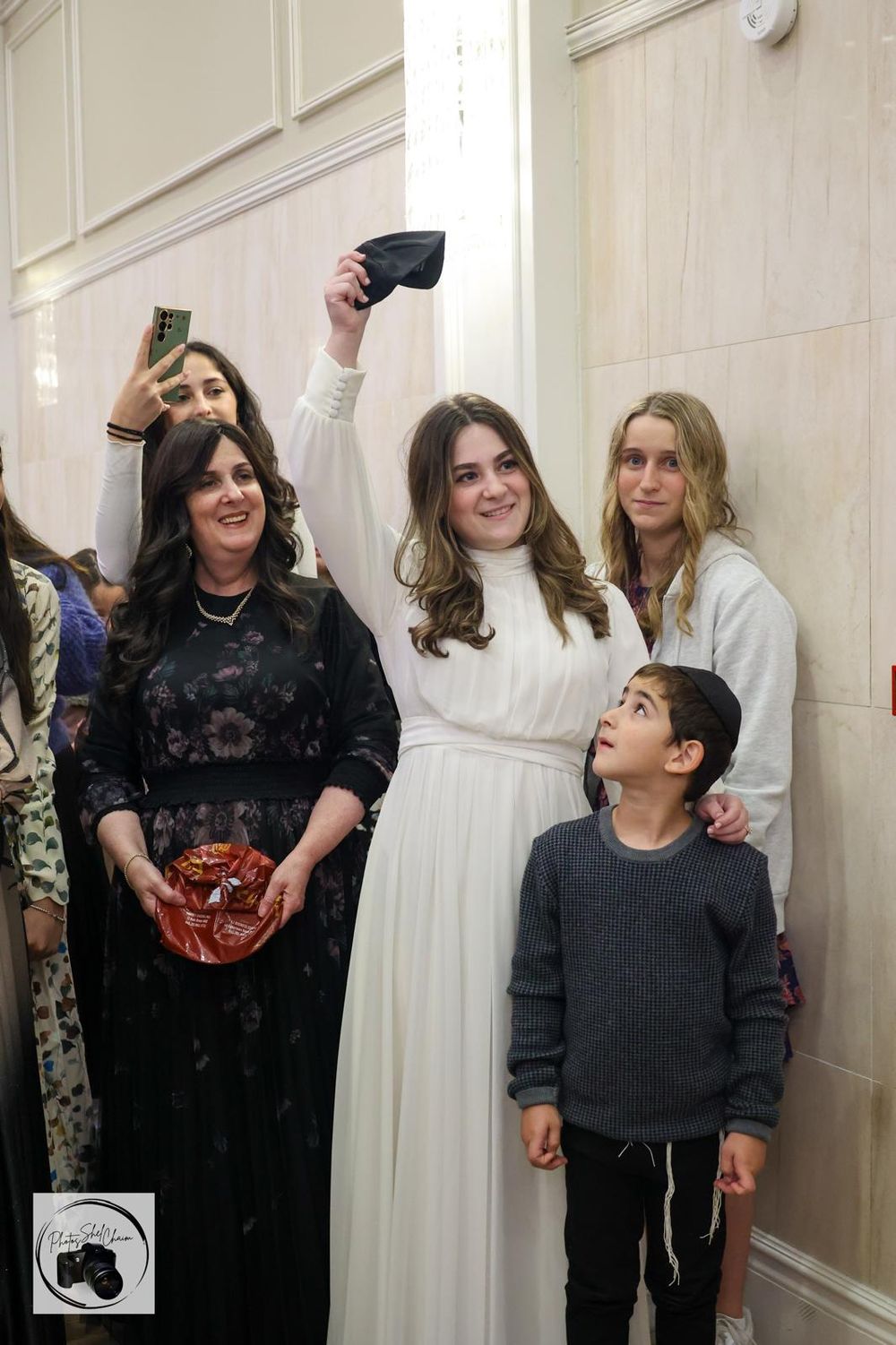 A woman in white dress takes selfie with family group including child and teen girls at formal indoor event.