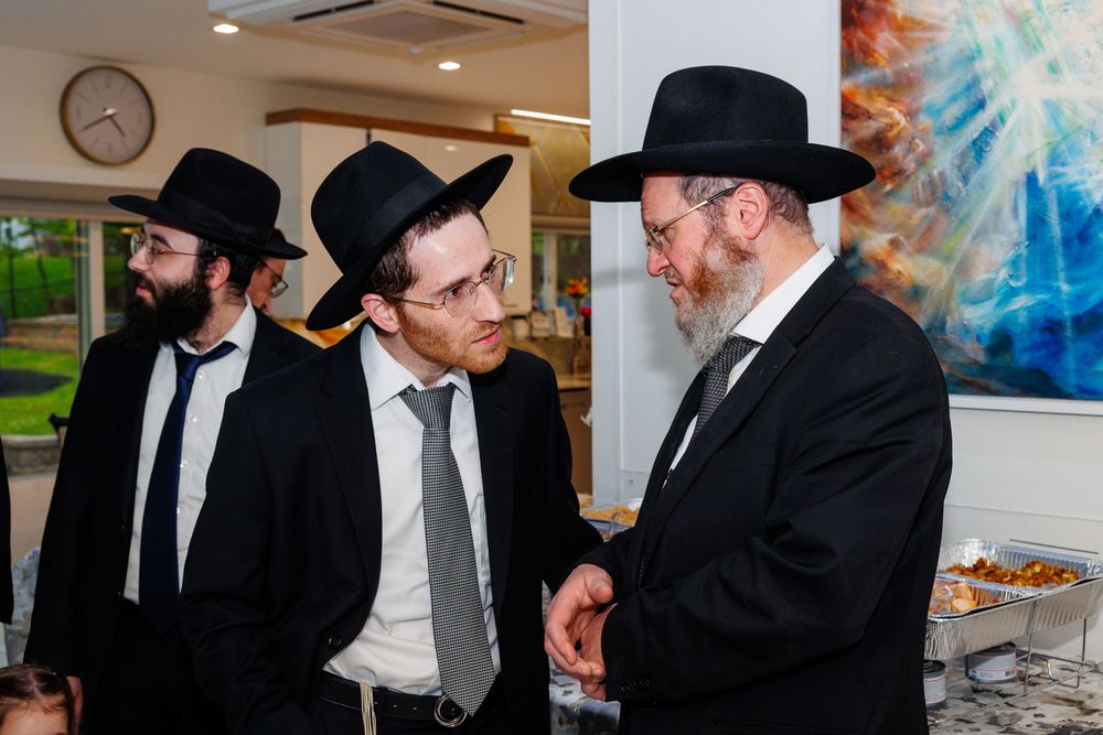 Orthodox Jewish men in black suits and hats engaged in conversation at a formal gathering or religious event.