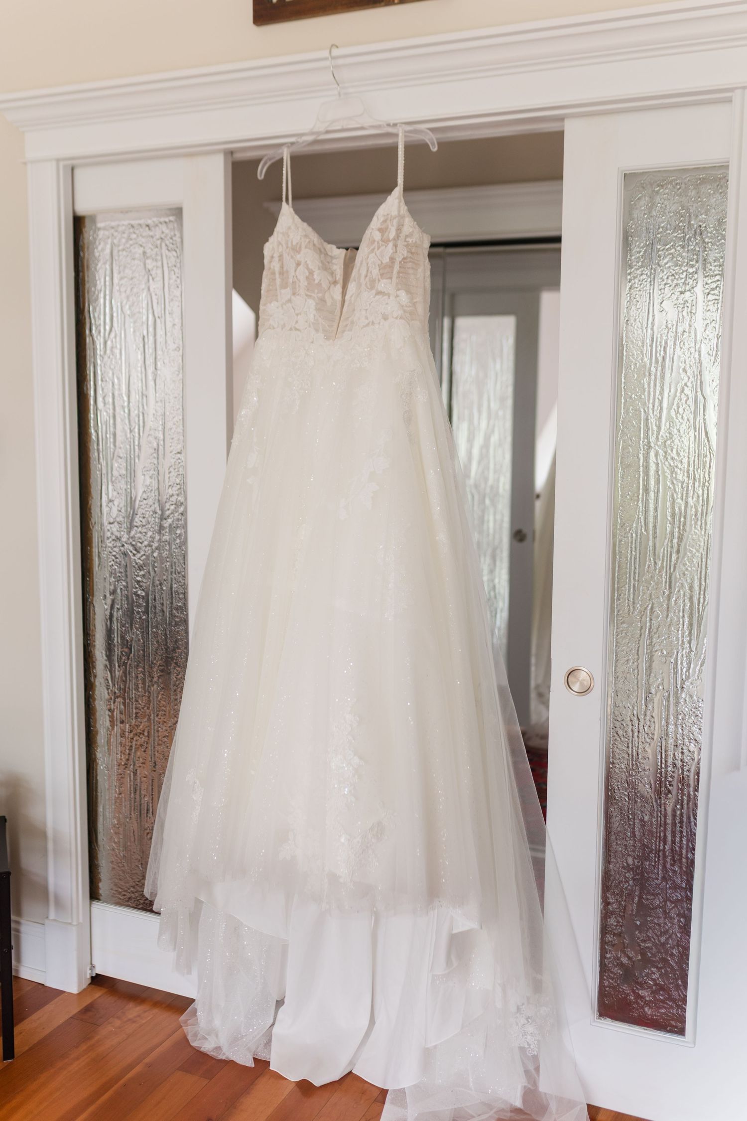 White lace A-line wedding dress hanging on a hanger between elegant frosted glass French doors.