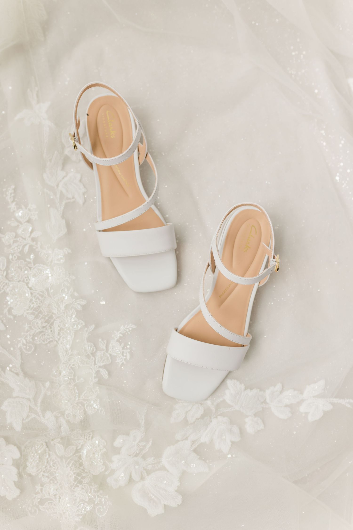 White bridal block heel sandals with ankle straps displayed on delicate lace wedding dress fabric.