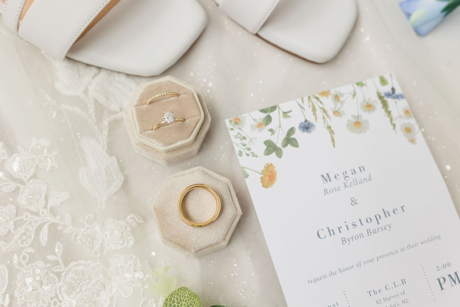 Wedding flat lay with floral invitation, diamond engagement ring, gold band in velvet box, and bridal heels on lace fabric.