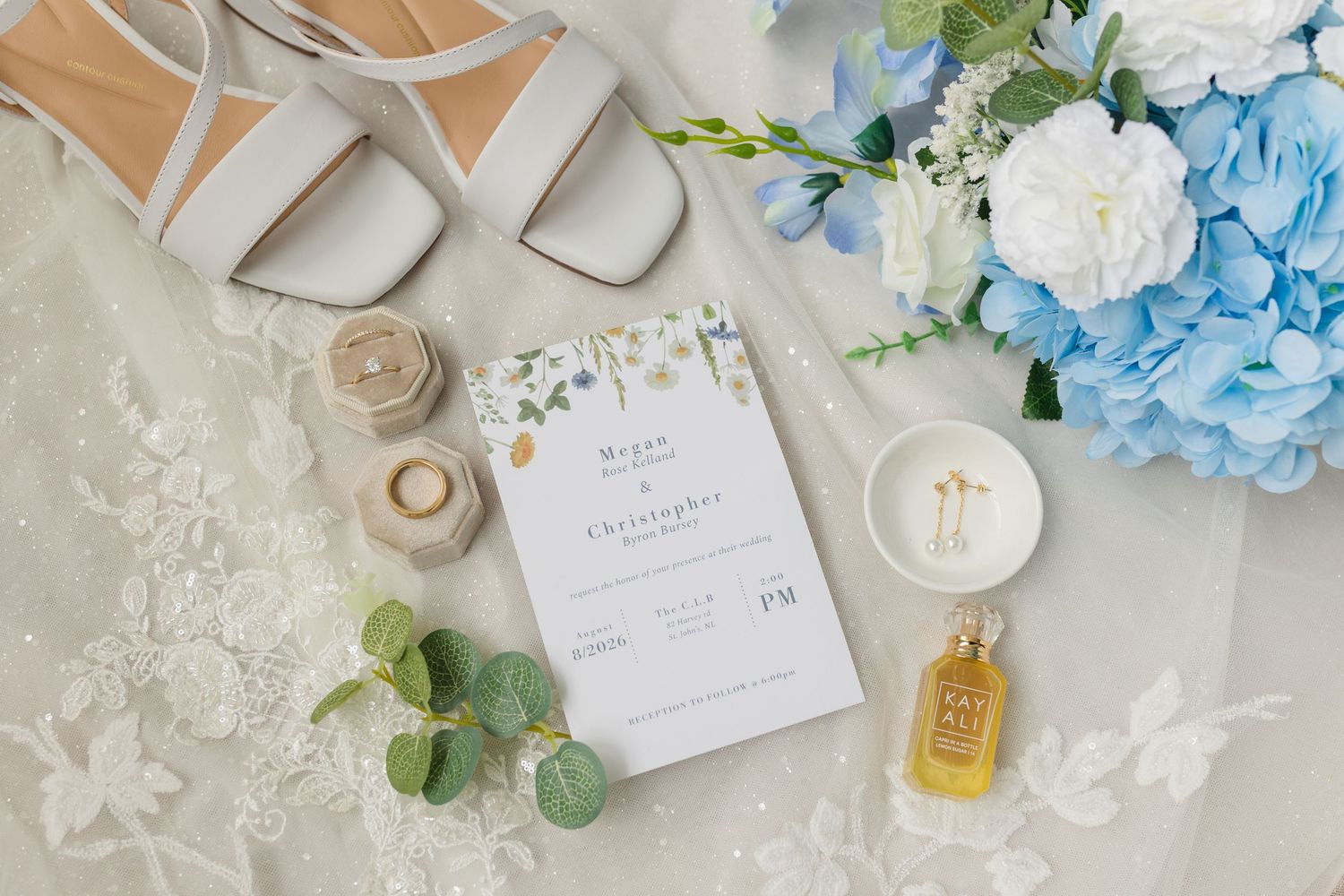 Elegant wedding flat lay with white heels, blue hydrangeas, floral invitation, ring boxes, and perfume on lace fabric.