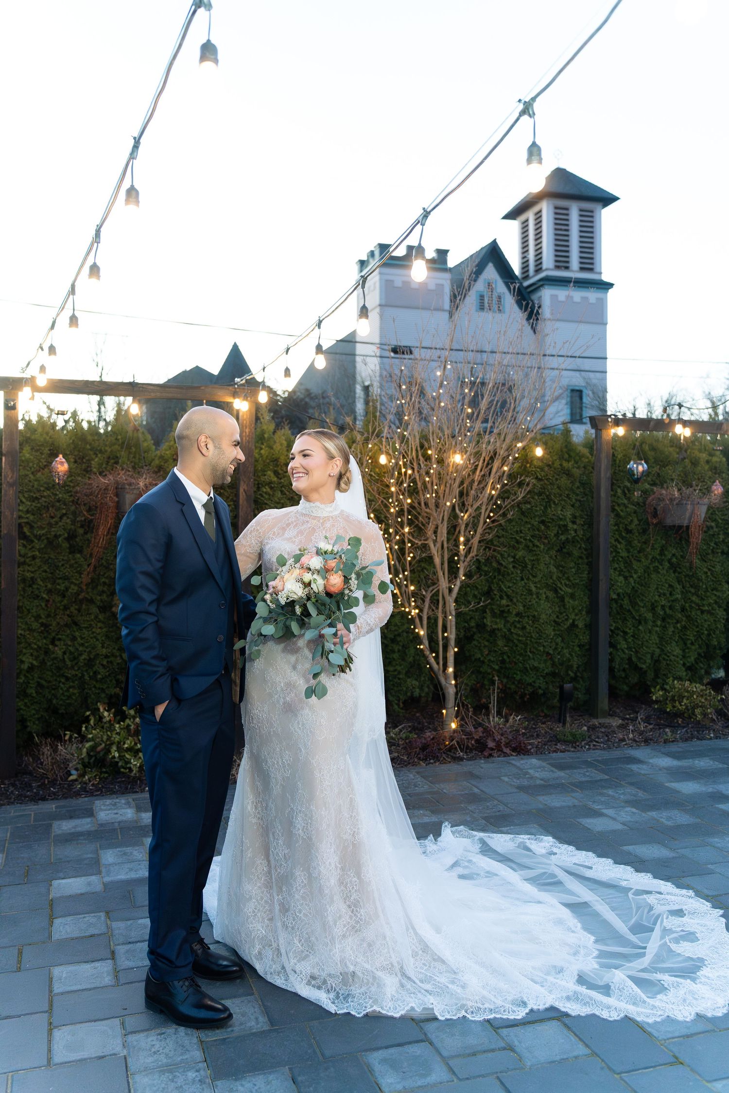 Bride and groom share a romantic moment on a patio with string lights and a church backdrop at dusk.