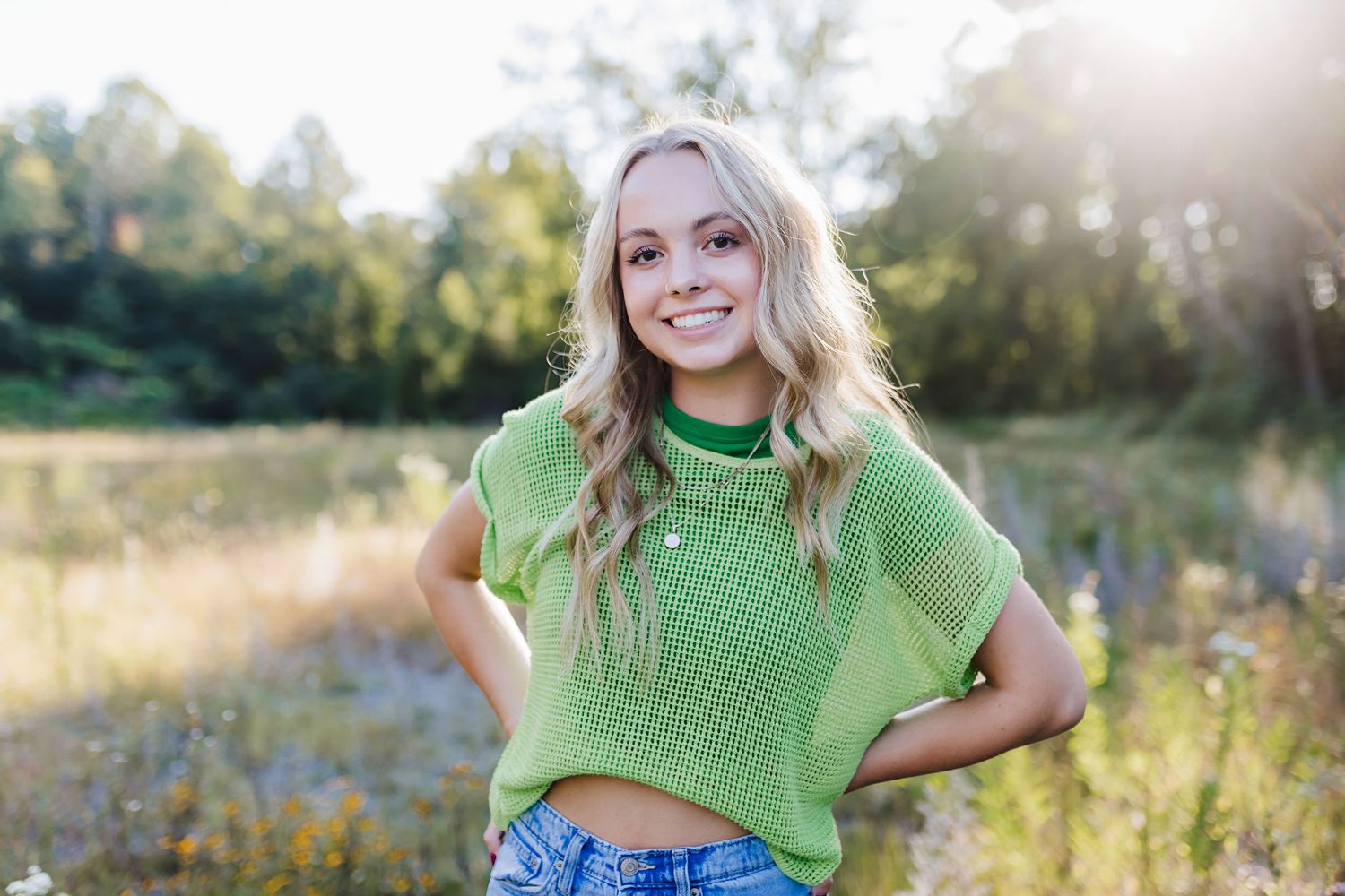 Smiling blonde teen girl in green knit top and denim shorts poses outdoors in sunlit field.