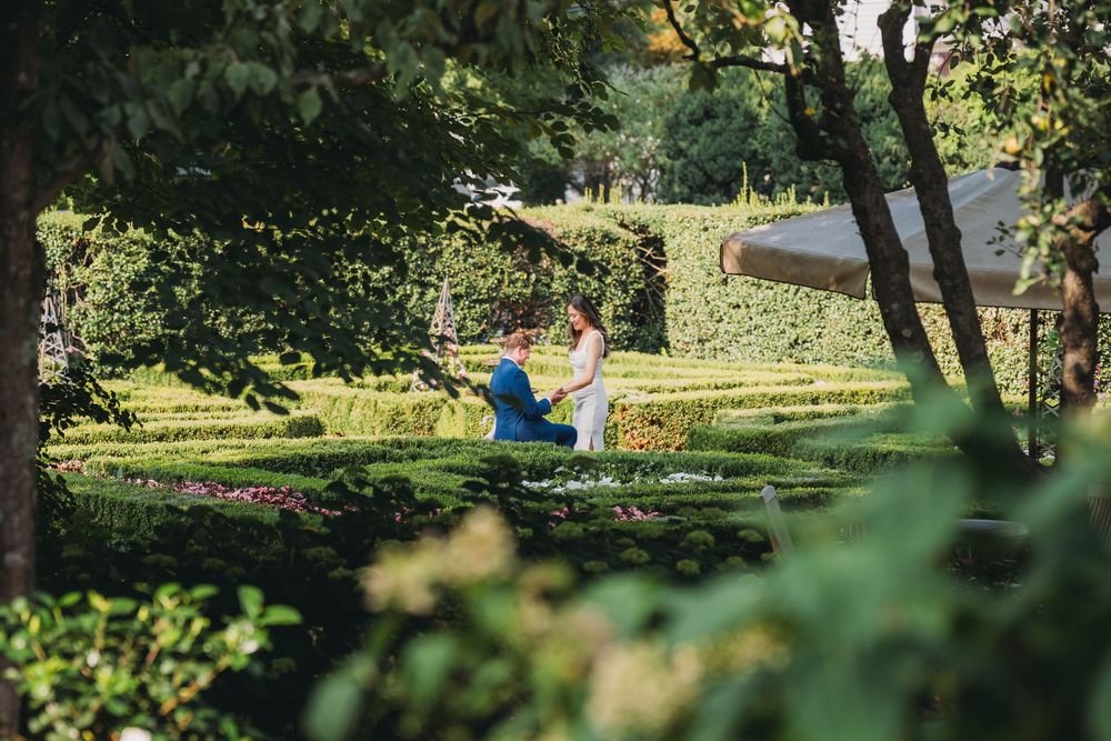 Couple sharing a romantic moment in a lush green garden with manicured hedges and colorful flowers.