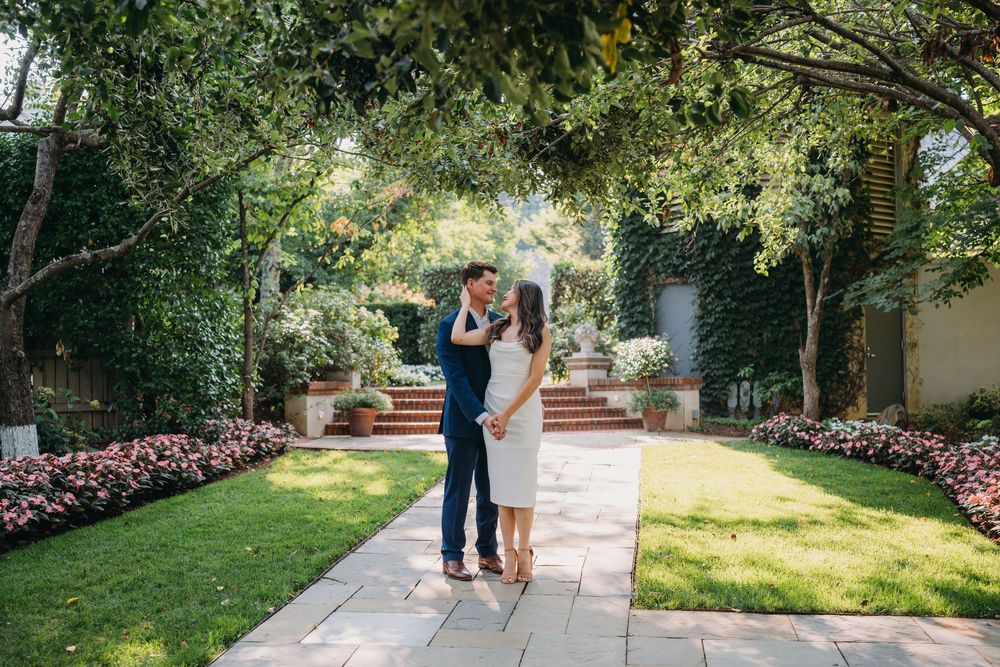 A couple shares a romantic embrace on a garden pathway surrounded by lush trees and colorful flower beds.