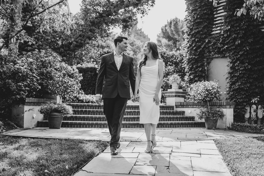 Black and white photo of a couple holding hands walking along a garden path surrounded by lush greenery and stone steps.