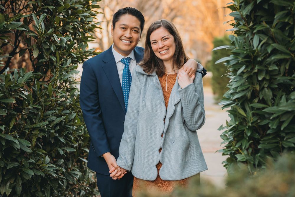 A smiling couple poses together outdoors among lush green hedges, man in suit and tie, woman in gray coat.