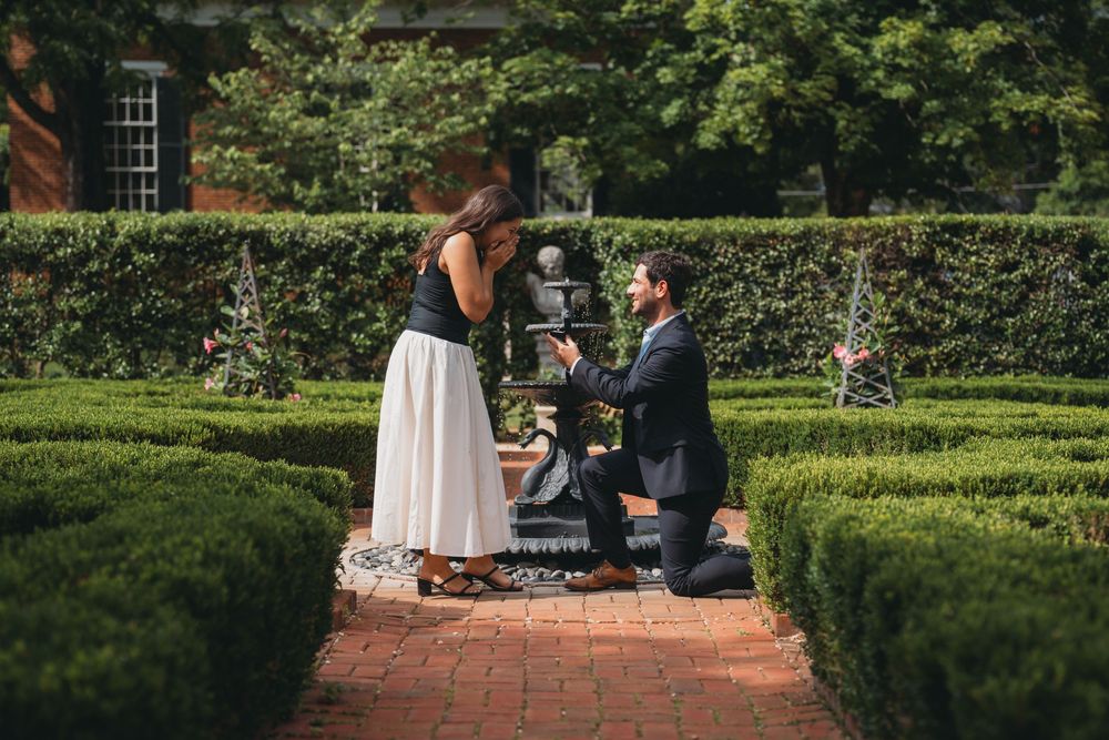 Man proposing to woman in a formal garden with manicured hedges on a brick pathway.