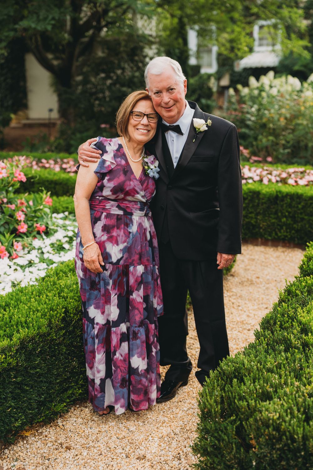Elegant couple posing in a garden, woman in floral gown and man in tuxedo with boutonniere, surrounded by manicured hedges.