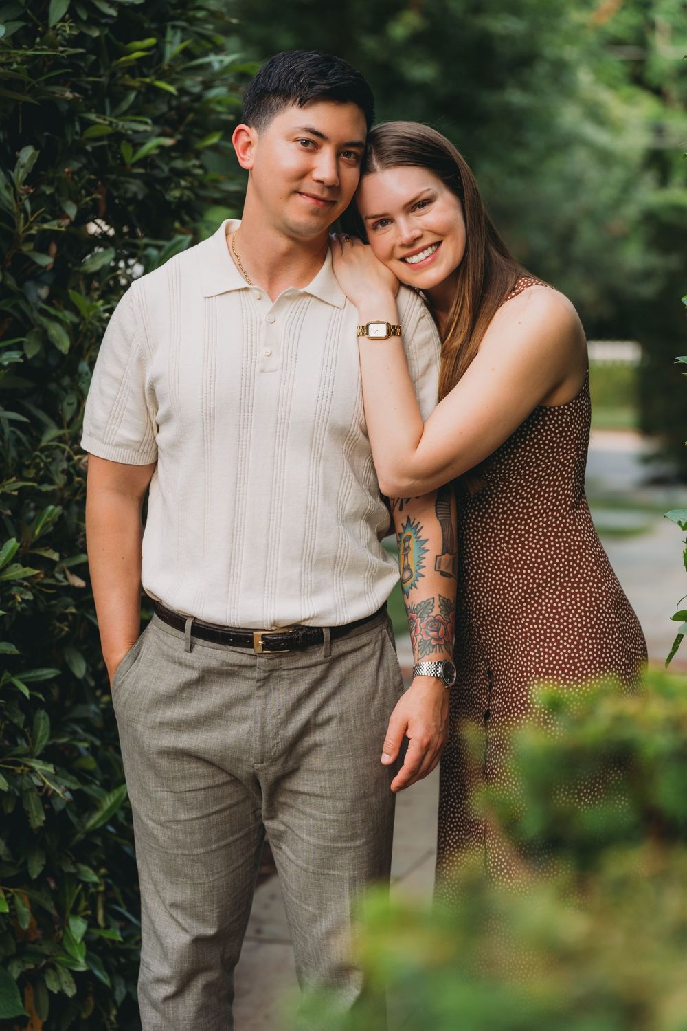 A smiling couple poses outdoors among green shrubs, woman with tattoos in floral dress leans on mans shoulder.