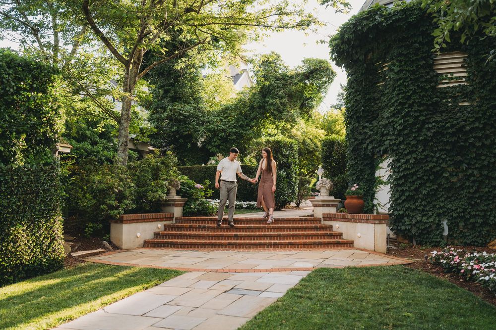 Couple holding hands walking through a lush garden with ivy-covered walls and brick steps.