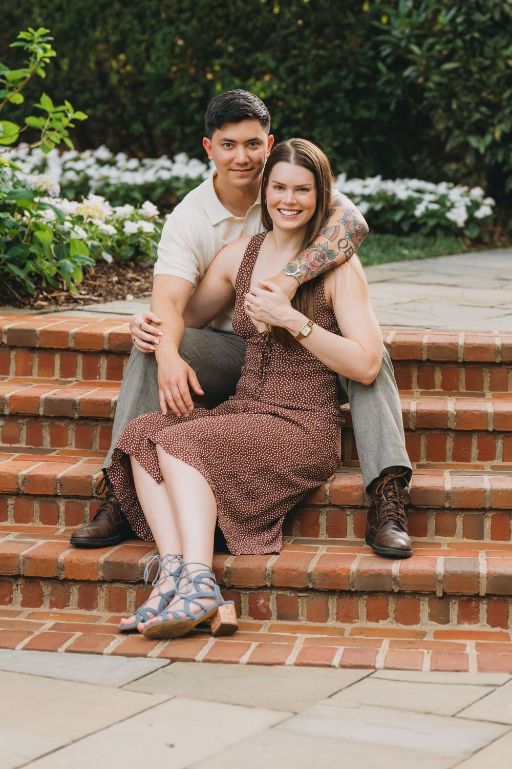 Couple sitting on brick steps in a garden, woman in floral dress with tattoos, man in beige polo and khakis.
