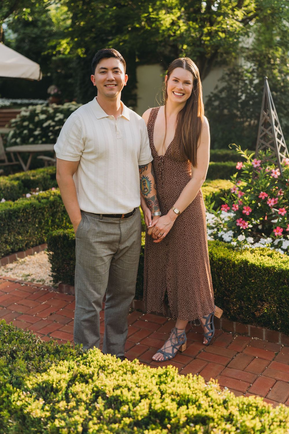 Smiling couple holding hands in a lush garden, man in white polo and gray pants, woman in brown floral midi dress.