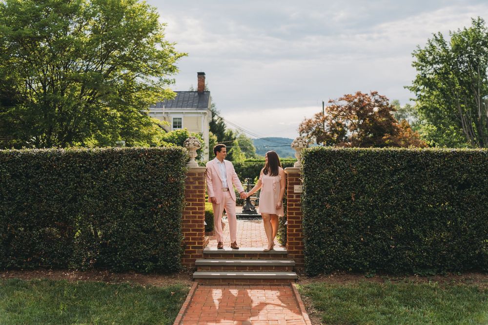 Couple holding hands walking through a hedge garden gateway toward a historic estate with mountain views.