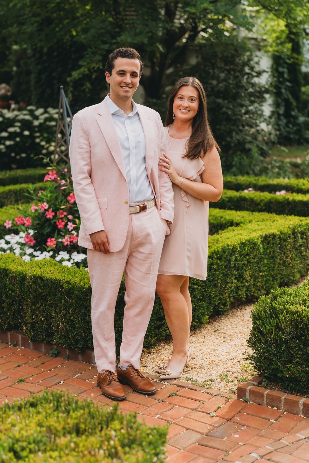 Couple in matching pink outfits posing in a formal garden with manicured hedges and colorful flowers.