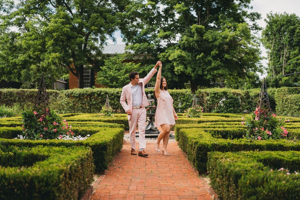 A couple dances joyfully in a formal garden with manicured hedges and brick pathways surrounded by lush greenery.