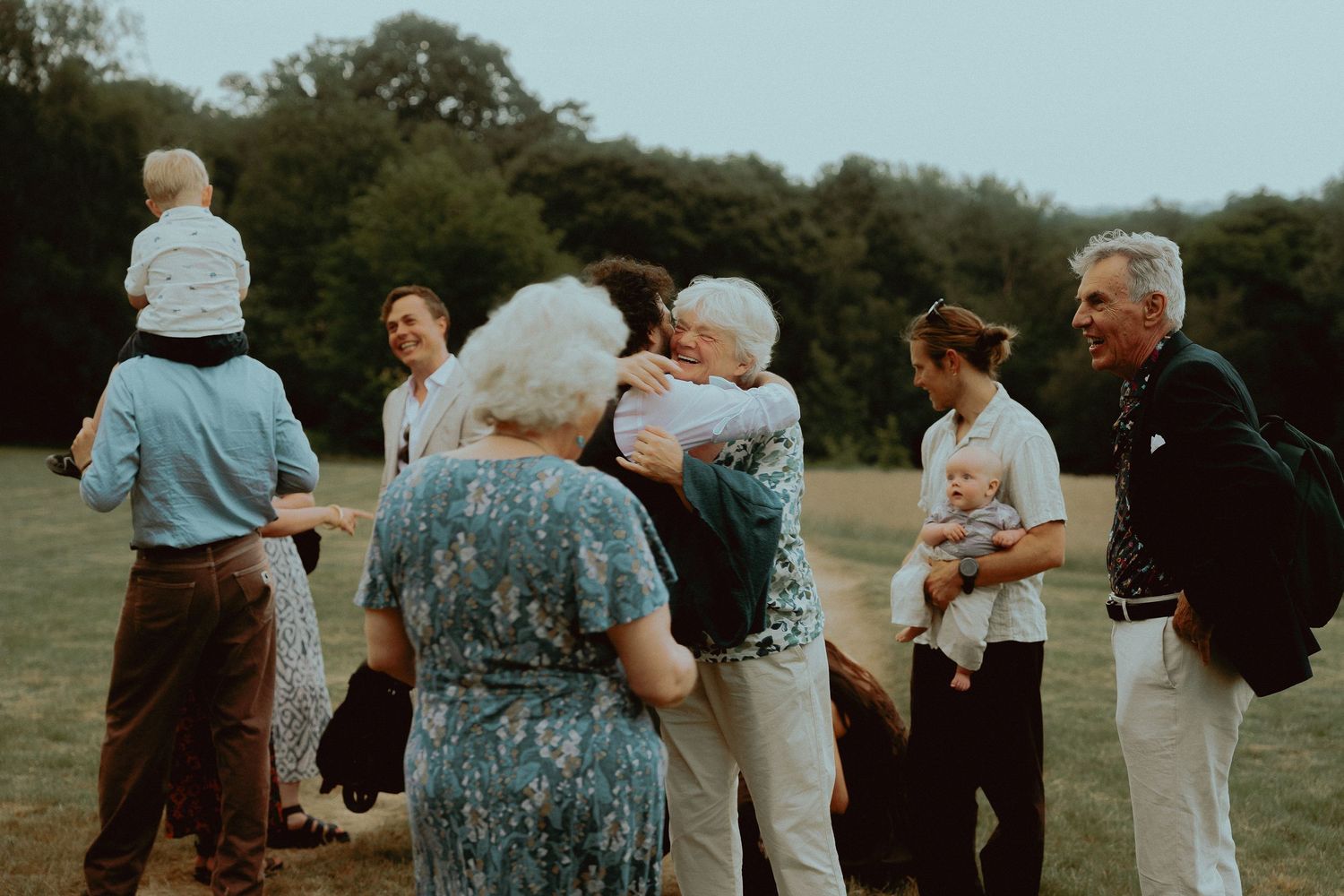 Family and friends gather outdoors, sharing warm hugs and laughter during a joyful reunion in a green park.