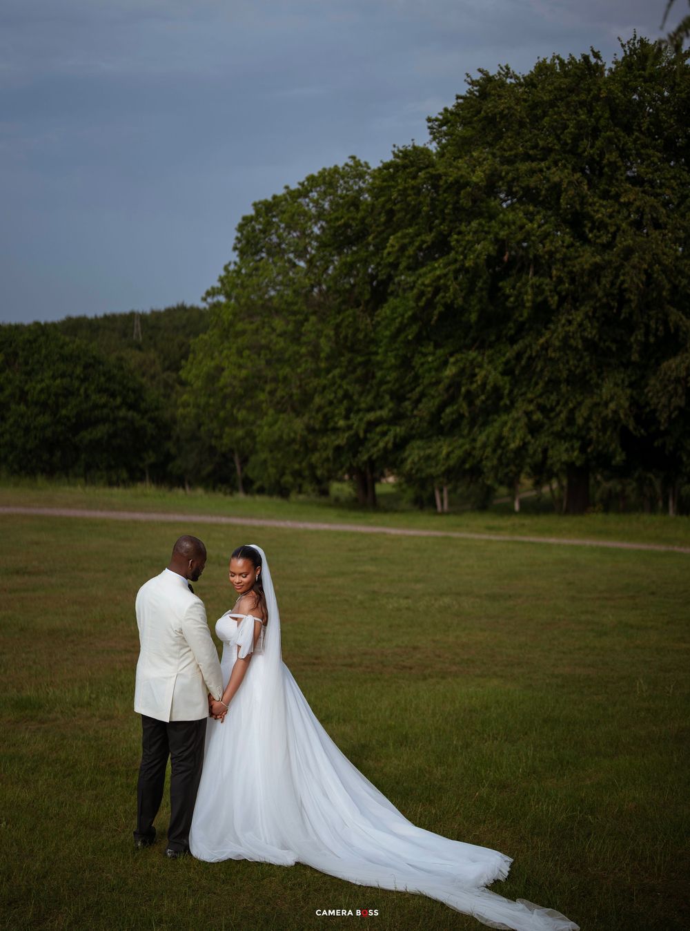 Bride in white gown with long veil holds hands with groom in white jacket on lush green lawn with trees.