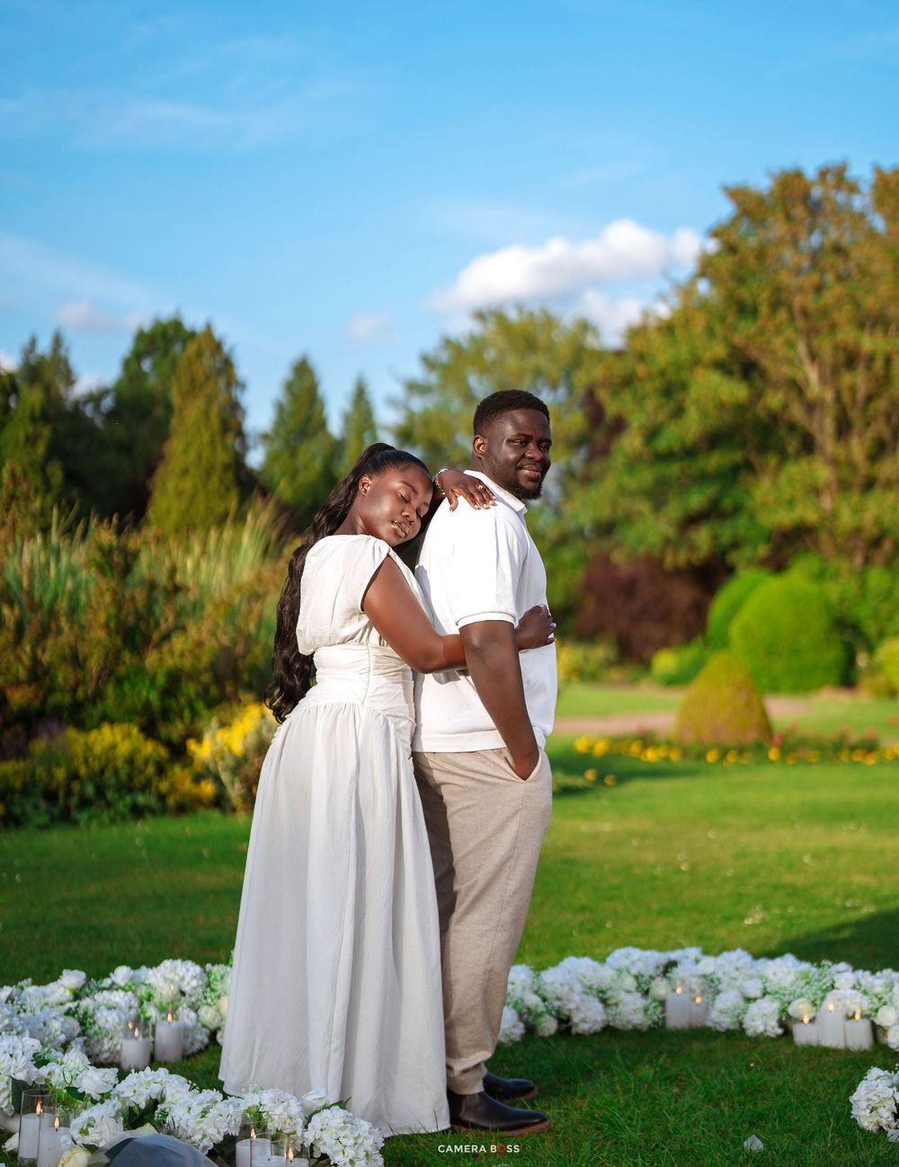 Couple in white outfits posing in a lush green garden surrounded by white flowers under a blue sky.