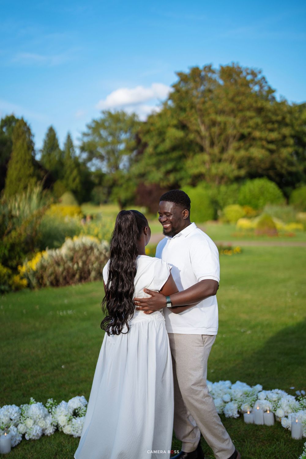 Couple in matching white outfits dancing outdoors surrounded by white flowers and candles in a lush green garden.
