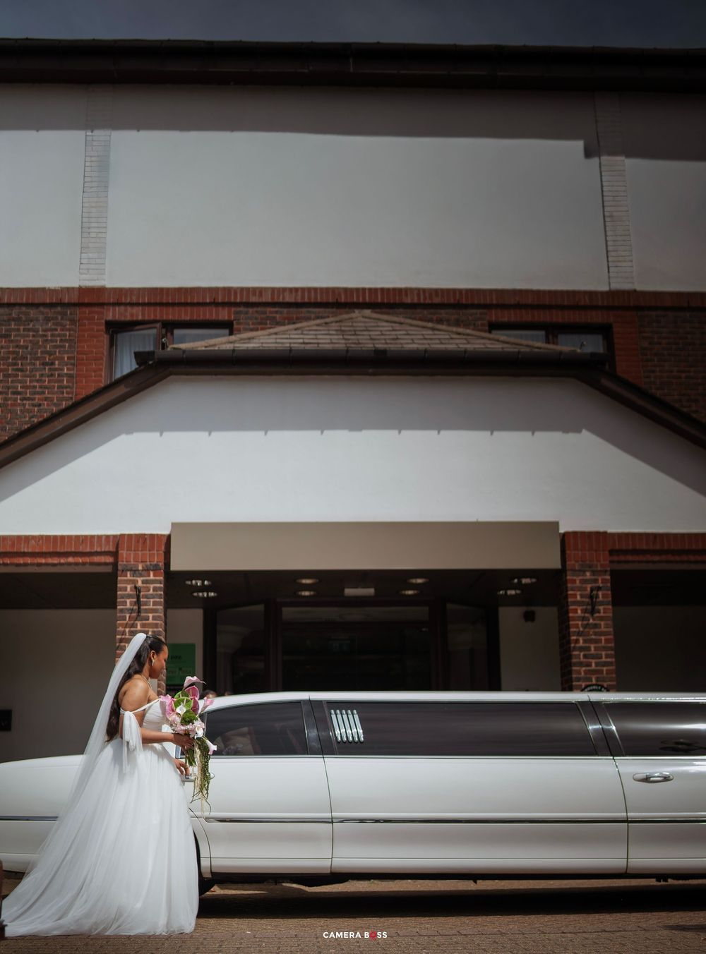 Bride in white gown and veil holding bouquet beside a white stretch limousine outside a brick building.