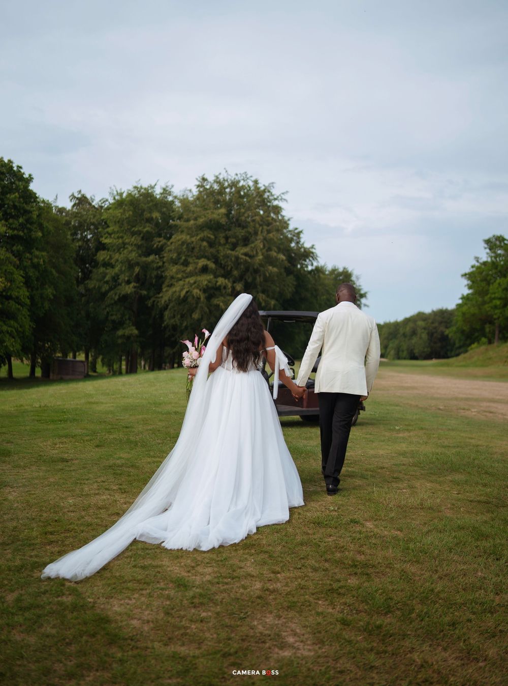 Bride in white gown with veil walks with groom in cream suit toward vintage car on lush green lawn.