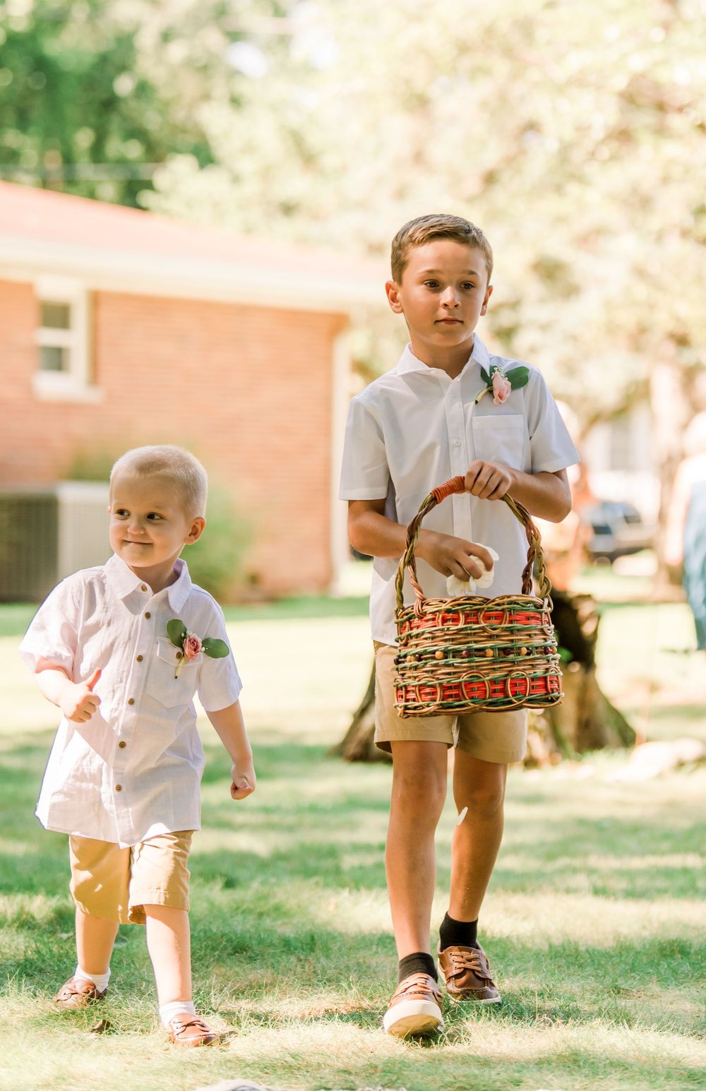 Two young ring bearers in white shirts and khaki shorts walk across green grass, one carrying a woven basket at an outdoor wedding.