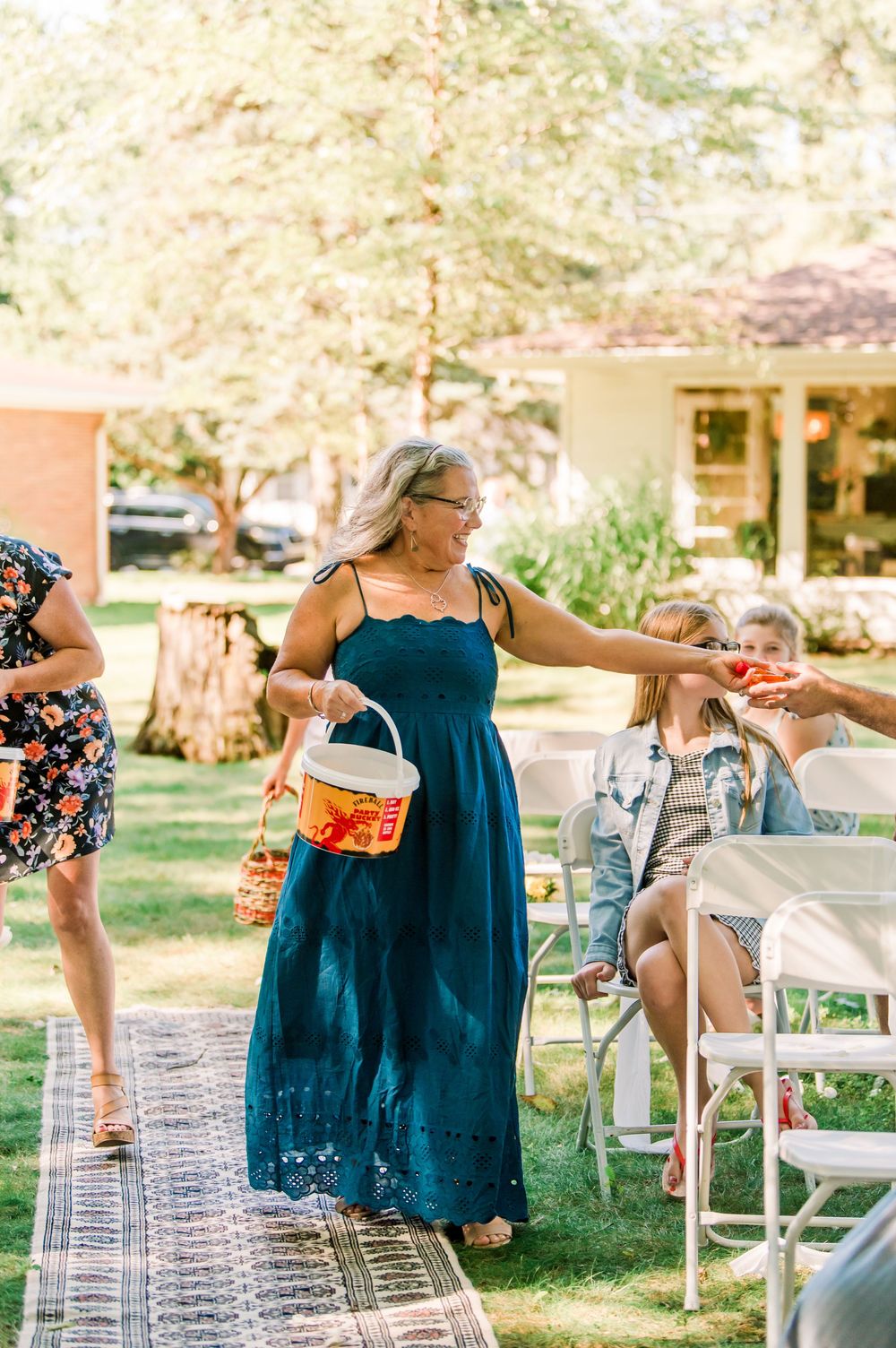 Woman in teal dress distributing items to guests at an outdoor wedding ceremony on a sunny day.