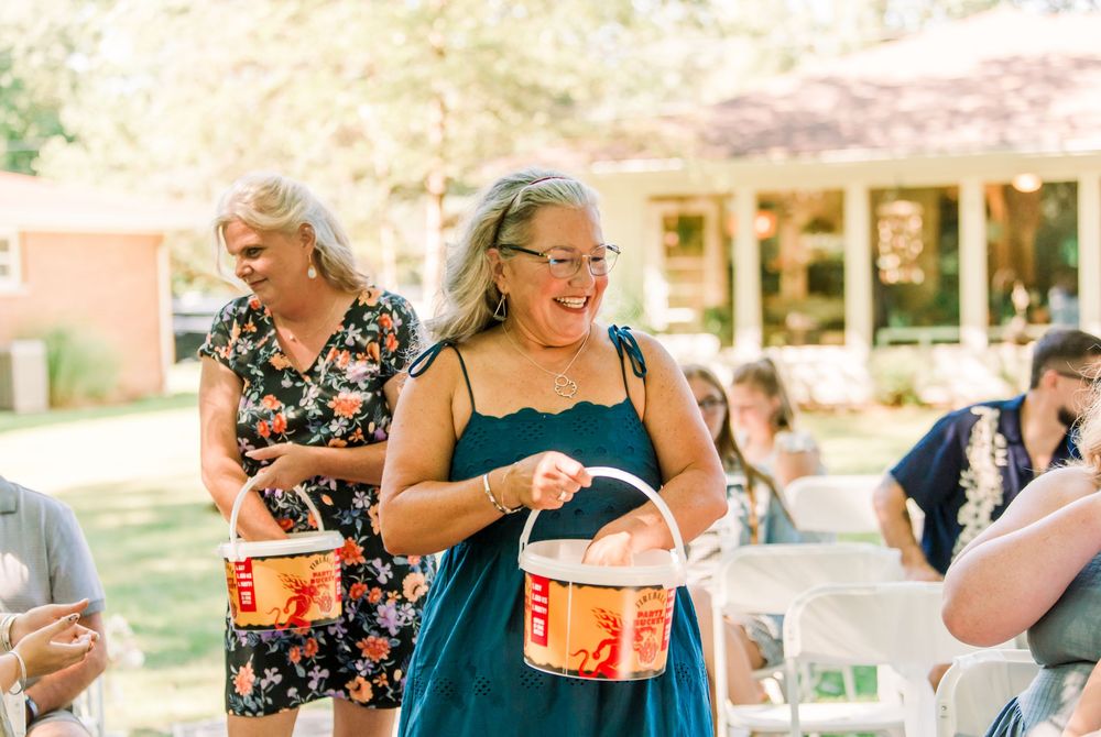 Woman in teal dress smiling while carrying orange bucket at outdoor summer gathering.