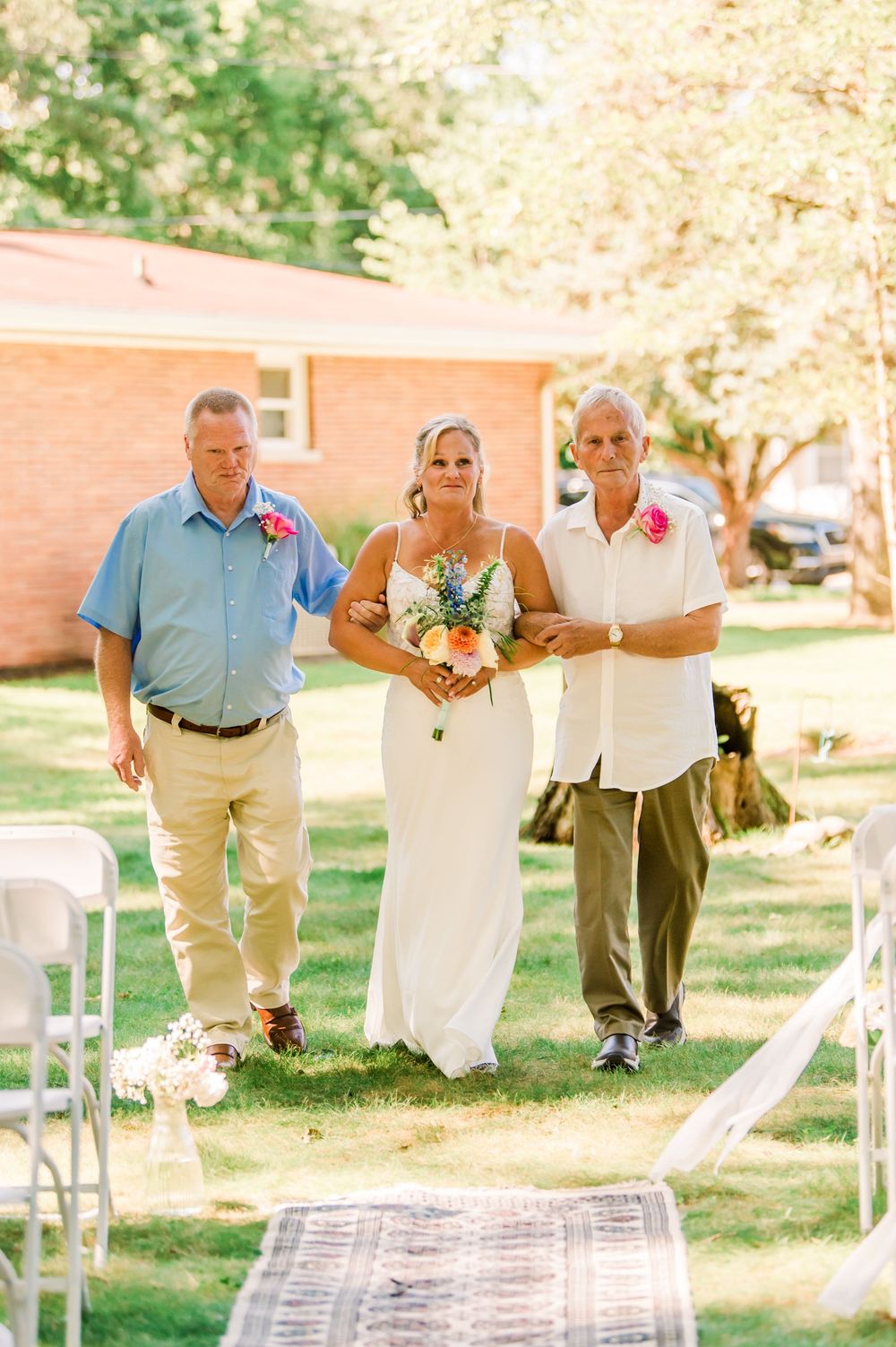 Bride walks down outdoor aisle escorted by two men in blue shirts, holding colorful bouquet at sunny garden wedding.