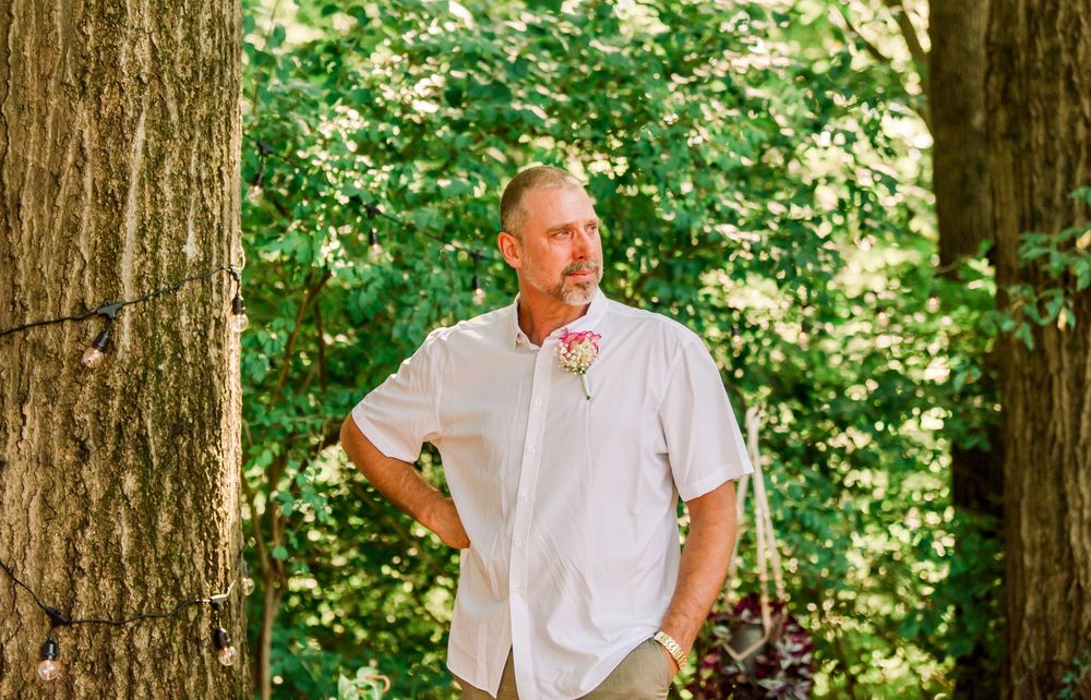 Man in white shirt with boutonniere standing among trees in a lush green forest setting.