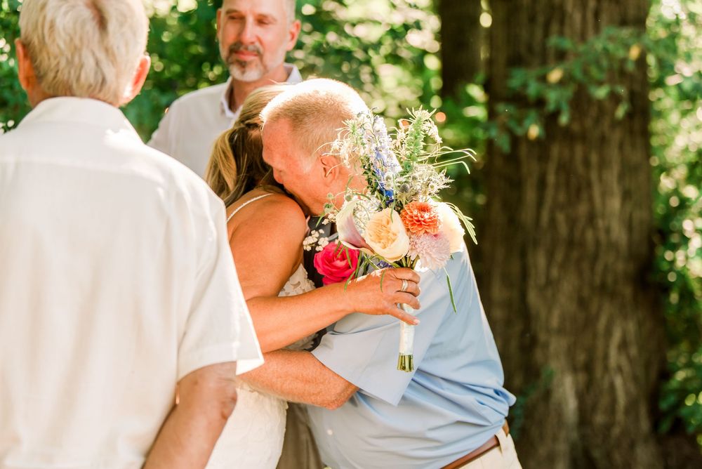 Bride hugging a guest while holding a colorful bouquet of flowers at an outdoor wedding ceremony.