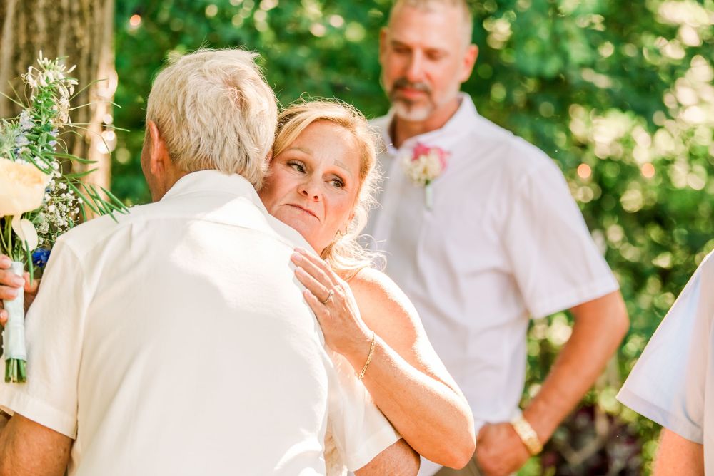 Bride embracing a guest at an outdoor wedding ceremony while the groom stands in the background among green trees.