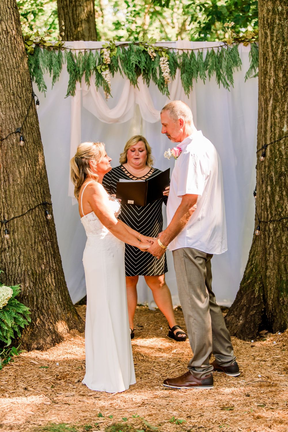Couple exchanging vows at outdoor wedding ceremony beneath rustic tree arch with white draping.