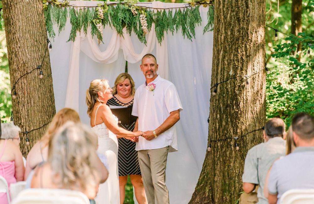 Smiling newlyweds holding hands with officiant at outdoor woodland wedding ceremony altar.