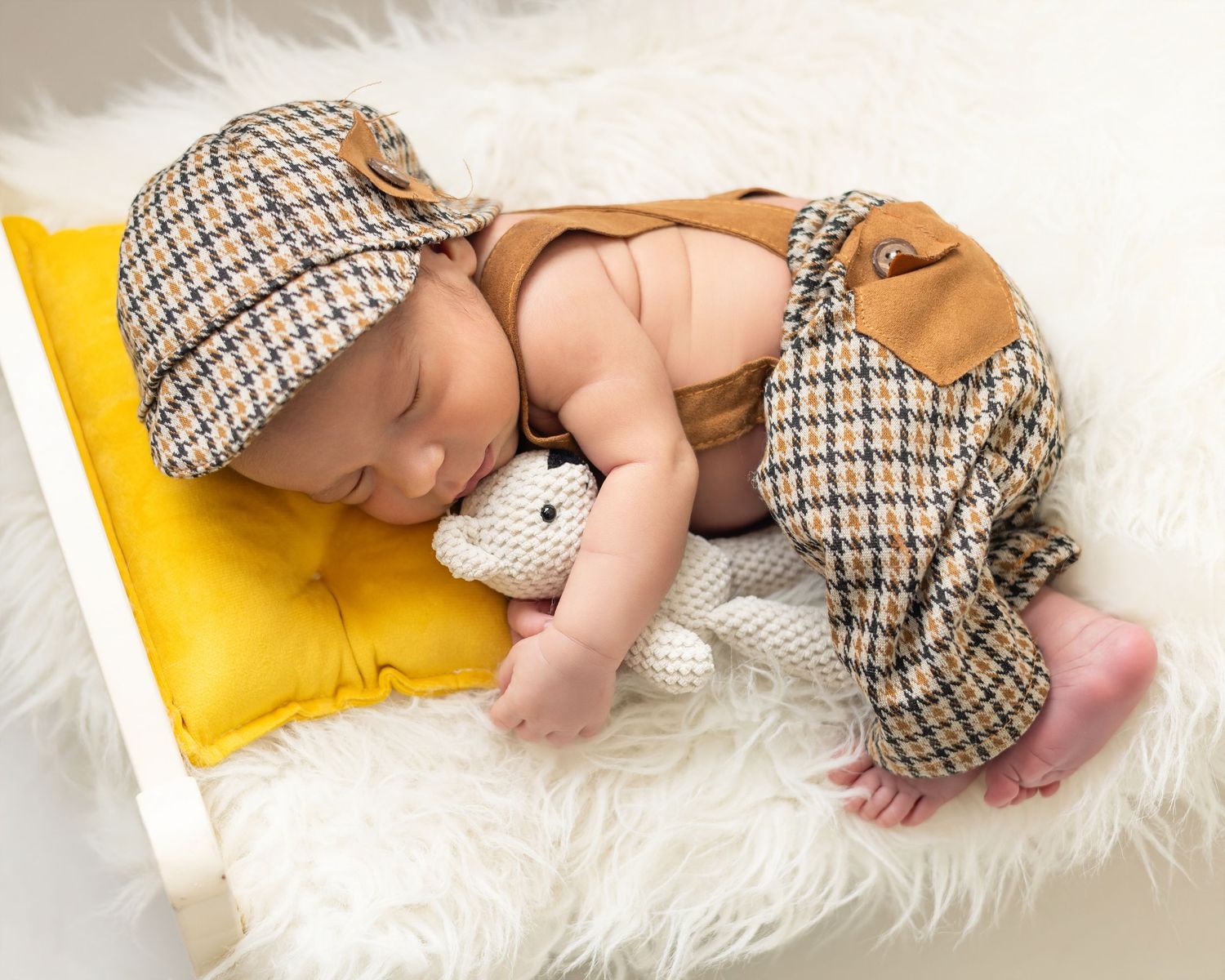 Sleeping newborn baby in houndstooth outfit and cap, resting on yellow pillow with stuffed toy on white fur blanket.