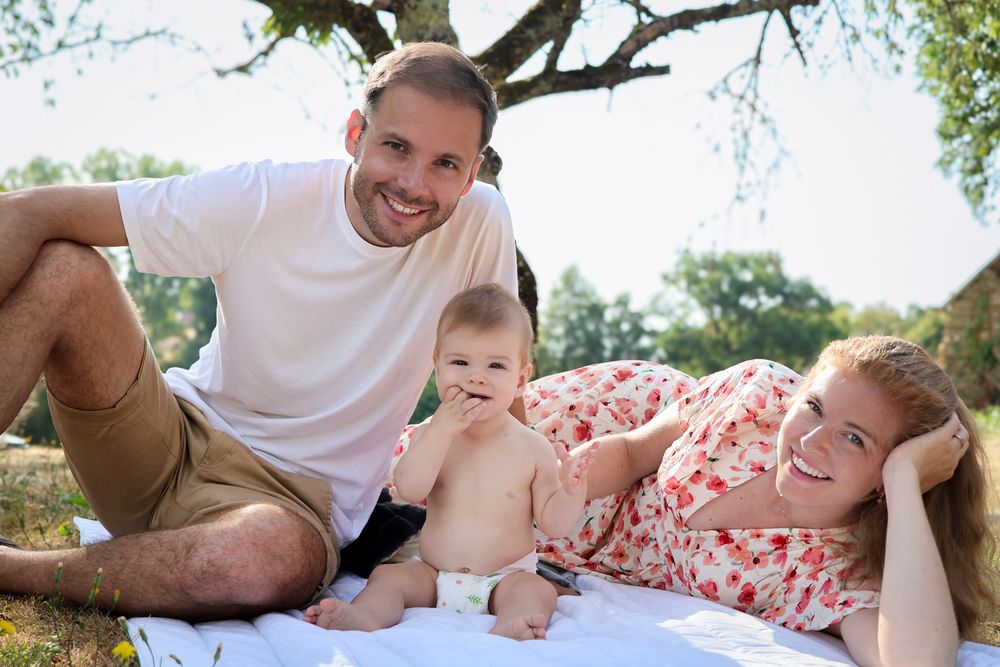 Père et mère souriants avec leur bébé sur une couverture à l’ombre d’un arbre, séance photo famille en Ardèche