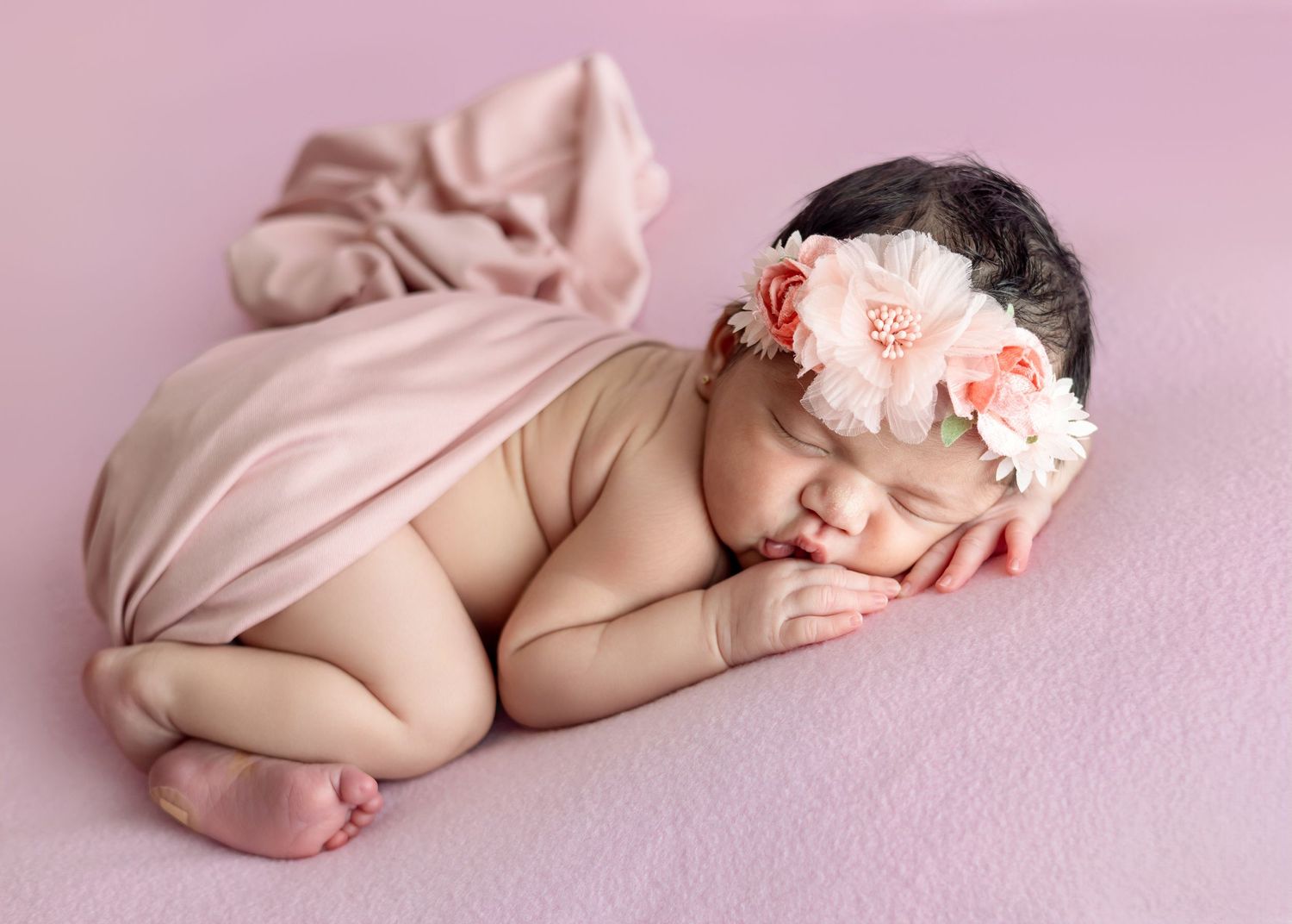 Newborn baby girl sleeping peacefully on pink blanket wearing a floral headband in a professional newborn photo.