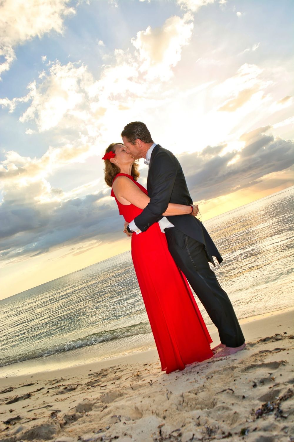 Couple sharing a romantic kiss on a sandy beach at sunset, woman in elegant red gown, man in dark suit.