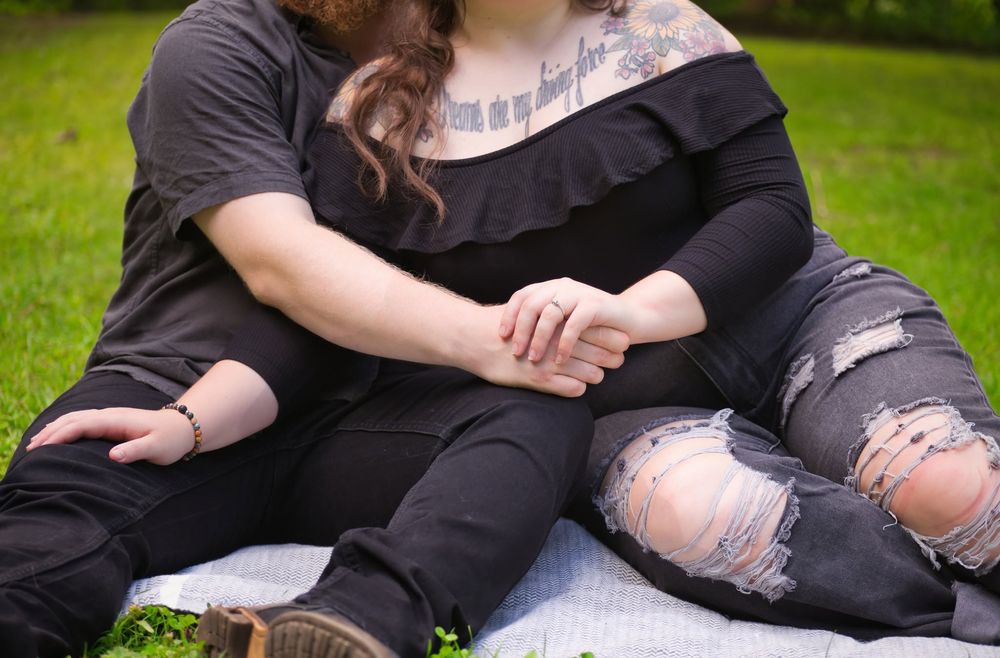Couple sitting on blanket in park holding hands, woman with tattoo wearing ripped black jeans.