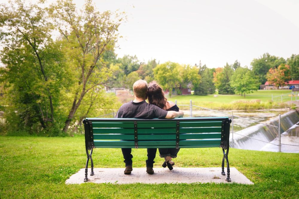 Couple sitting closely together on a green park bench, enjoying a peaceful view of a river and lush trees.