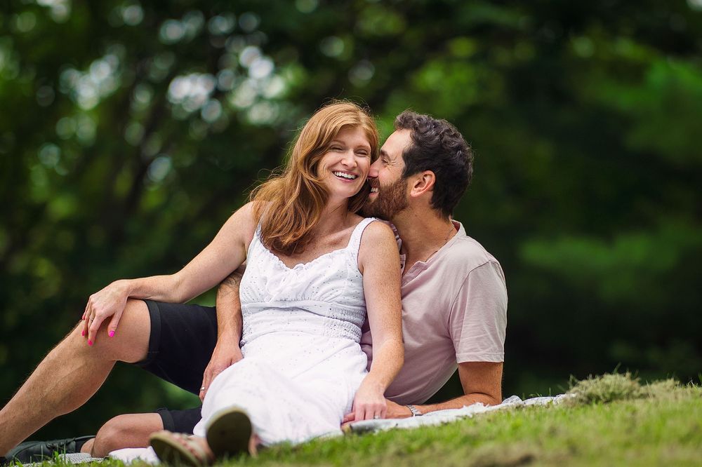 A couple sits on grass in a park, sharing a tender moment as the man leans in to kiss the woman's cheek.